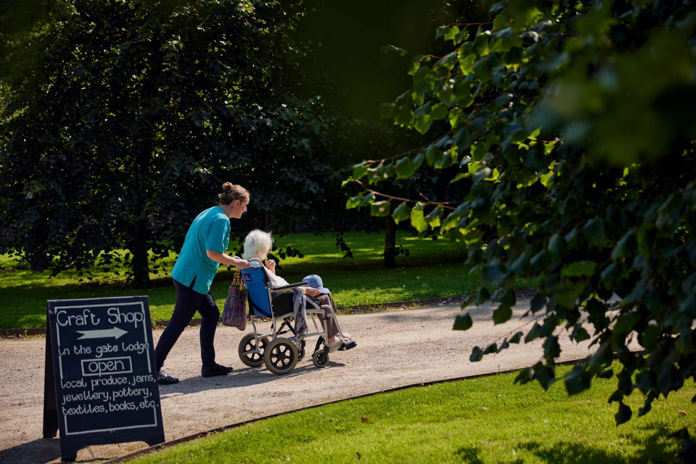 nursing home worker in Borris Lodge Nursing Home wheeling a resident along a wooded path with a sign for a cafe